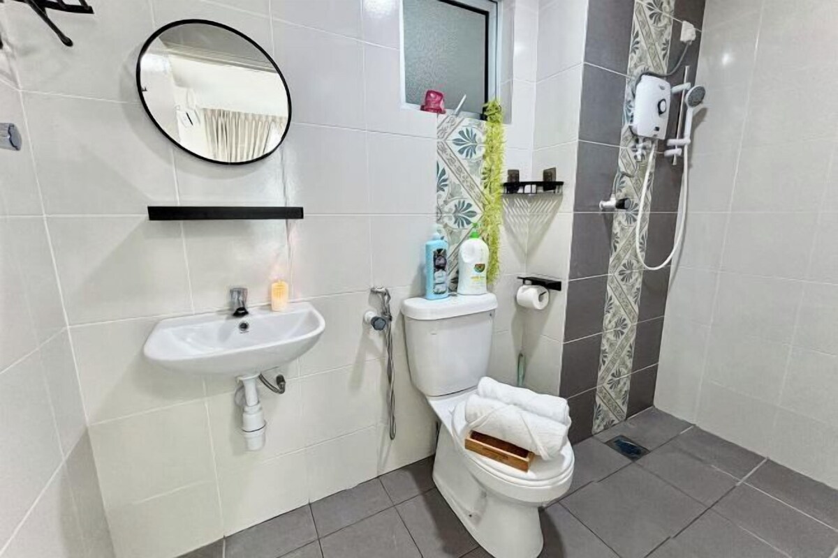 Une salle de bain avec un lavabo, des toilettes et une douche, tous de couleur blanche. Un miroir est placé au-dessus du lavabo et un porte-serviettes se trouve à côté des toilettes.