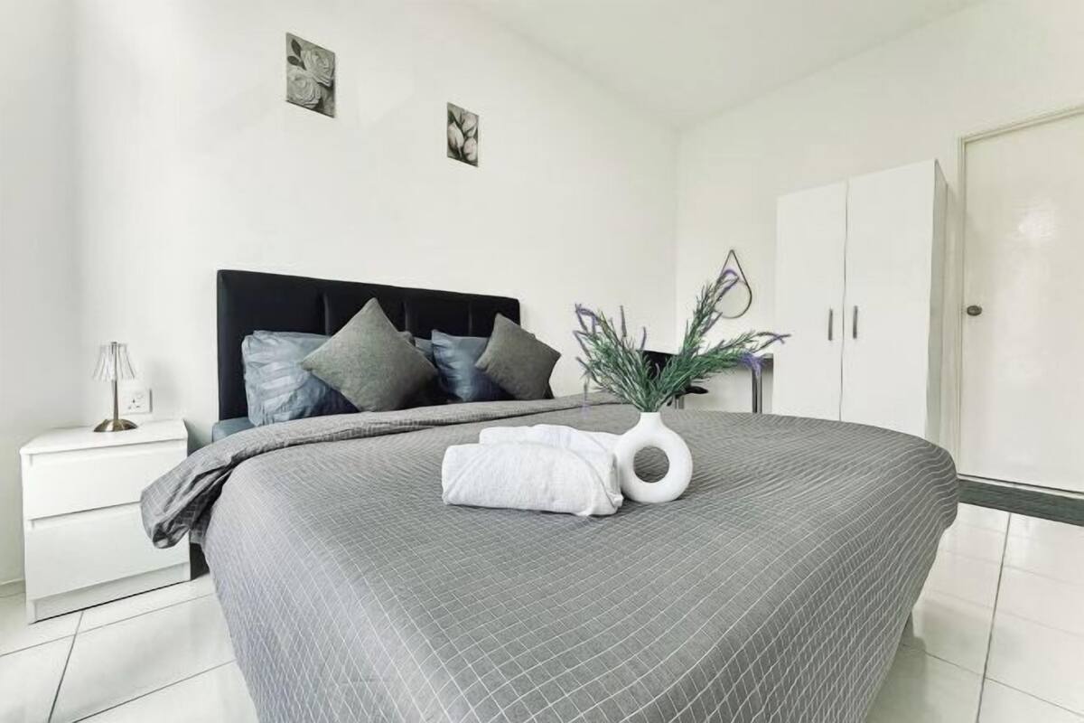Un lit bien fait, recouvert d’une couette grise, une table de chevet blanche avec une lampe, et une armoire blanche.