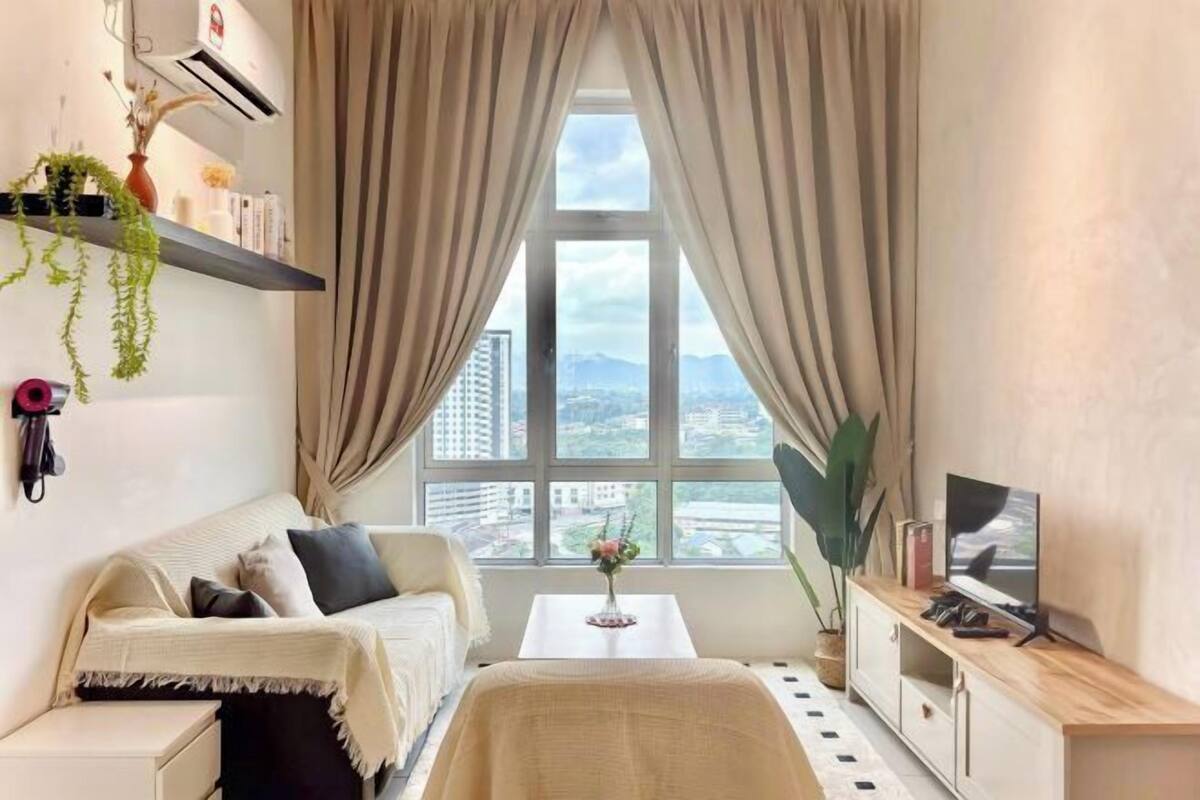 Une chambre à coucher avec un lit, une télévision, une fenêtre avec des rideaux, une petite table et une étagère avec des plantes.