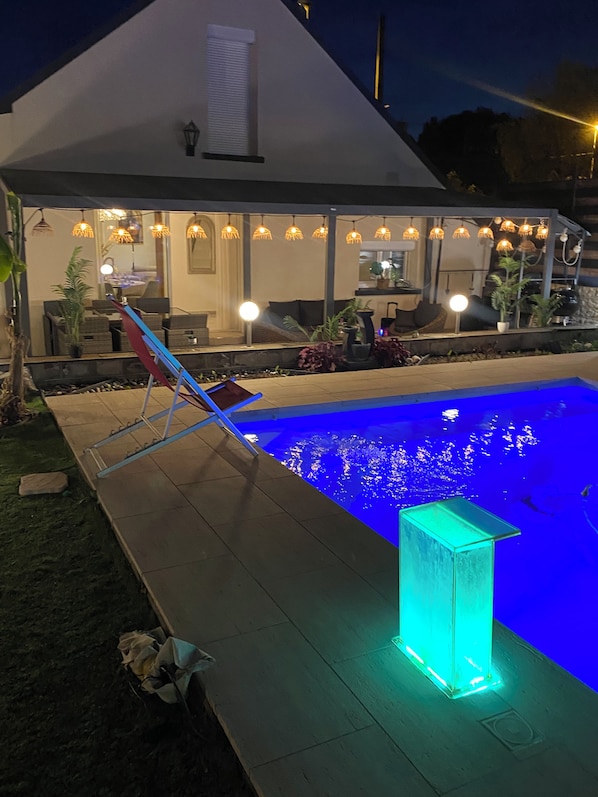 Ein Schwimmbad mit blauem Licht, eine überdachte Sitzgruppe mit hängenden Lampen und ein Blumentopf.