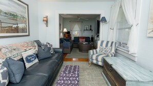 Ein Wohnzimmer mit einem dunklen Ledersofa, einem gemusterten Ottomanen und einem hellblauen Sessel. Eine Holzbank steht an der Wand, und über dem Sofa hängt ein gerahmtes Gemälde.