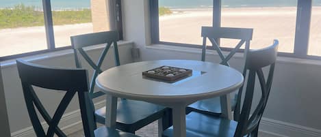 A dining area with a round table and chairs, large windows with blinds, and a view of the beach.