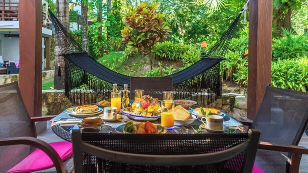 A dining table set with breakfast items, surrounded by chairs, in a lush garden setting with a hammock and wooden pillars. - Tropical Oasis w/ Cenote & Pool – Walk to Beach (Playa del Carmen)