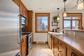 A kitchen with wooden cabinets, stainless steel appliances, a window with a view of snow-covered trees, and a hanging light fixture. - Tremblant Prestige - Altitude 172-11 (Mont-Tremblant)