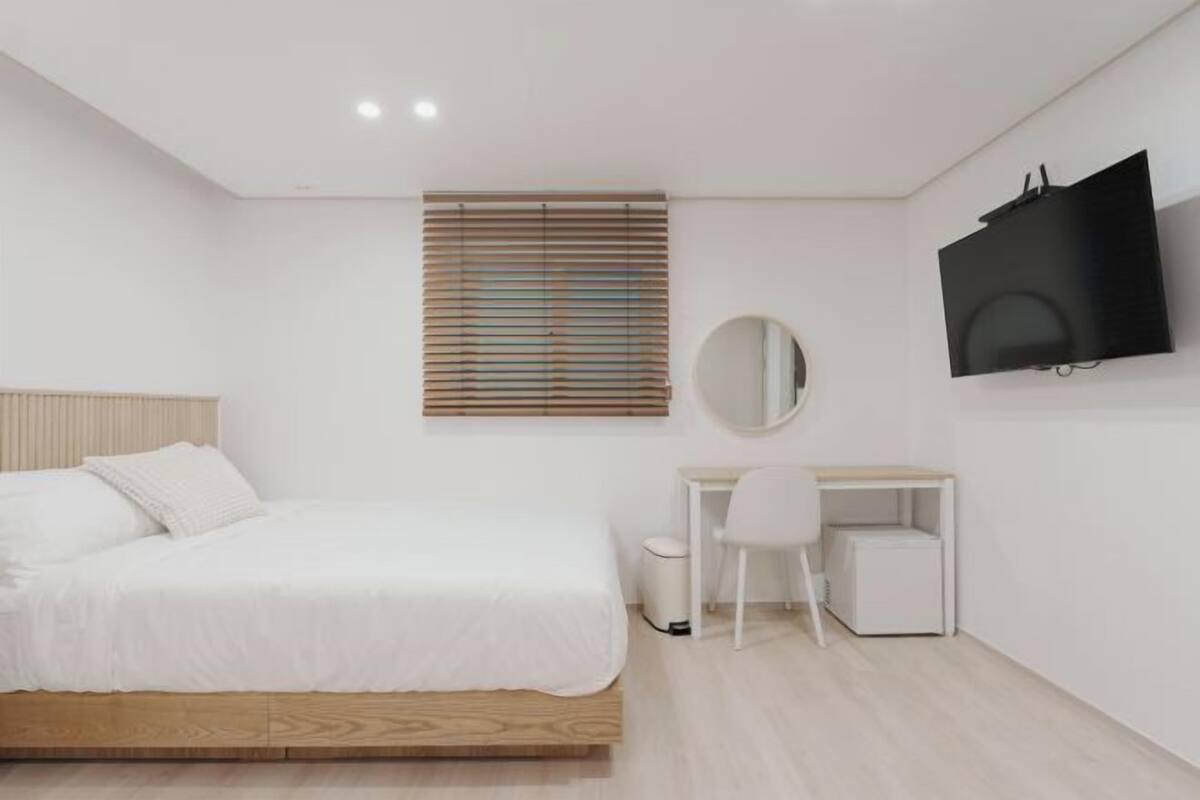 A bedroom with a bed, a desk, a chair, a television, and a window with blinds.