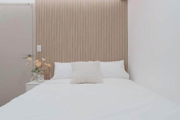 A neatly made bed with white linens and a single decorative pillow. A small bedside table holds a clock and a vase with flowers. The room features a vertical striped wall panel and a plain white door.