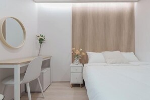 Um quarto moderno com cabeceira de madeira, mesa de cabeceira com relógio, escrivaninha branca com cadeira e espelho na parede.