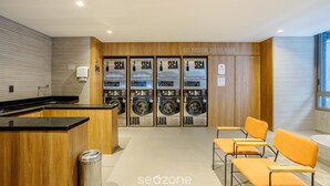 Un espace buanderie équipé de quatre machines à laver SECA, d’une poubelle et d’un coin salon avec des chaises orange.