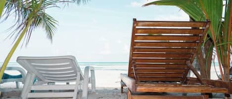 A wooden beach chair and a white plastic chair under a palm tree on a sandy beach with the ocean in the background.