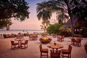 Un espacio exterior para comer con mesas y sillas, un hogar para fuego y vistas a la playa y al océano.