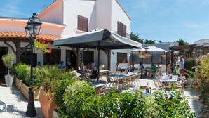 An outdoor café with tables and chairs, surrounded by greenery and a white building with a tiled roof.