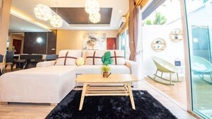 A modern living room with a sectional sofa, a coffee table, and a black furry rug. There is a dining area in the background with a table and chairs. - Aurora Life Pool Villa Pattaya (Pattaya)
