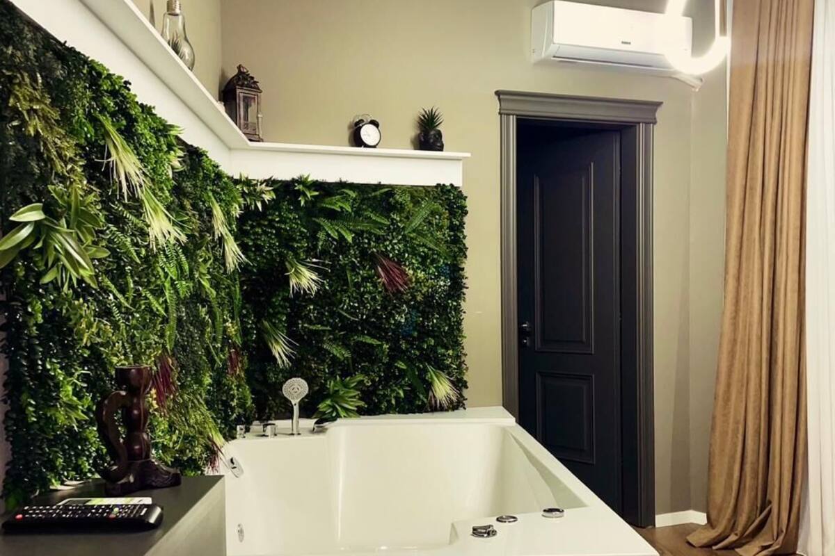 Ein modernes Badezimmer mit einer großen Badewanne, einer grünen Wand mit Pflanzen, einer kleinen Ablagefläche mit Dekoration und einer Tür mit Vorhang.