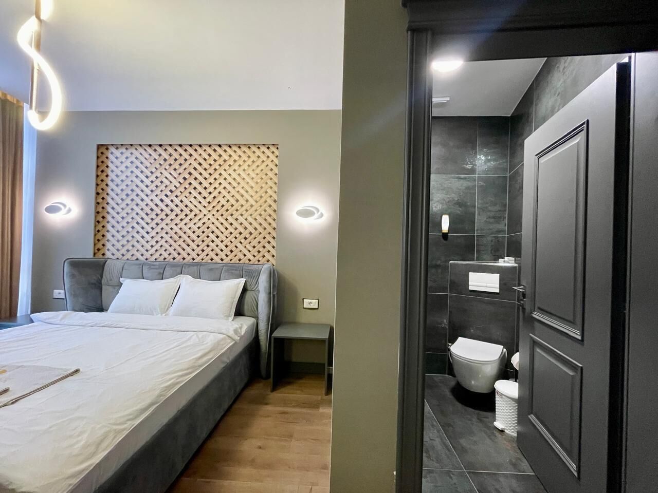 A modern bedroom with a bed, bedside table, and a patterned headboard. Adjacent is a bathroom with a toilet, sink, and mirror.