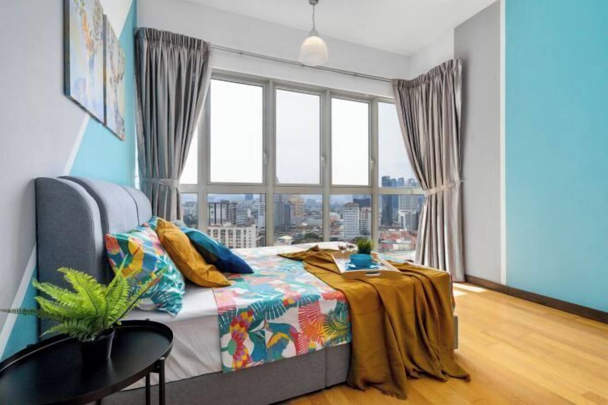Ein modernes Schlafzimmer mit großem Fenster, einem Bett mit bunter Bettwäsche, einer Pflanze und Blick auf die Stadt.
