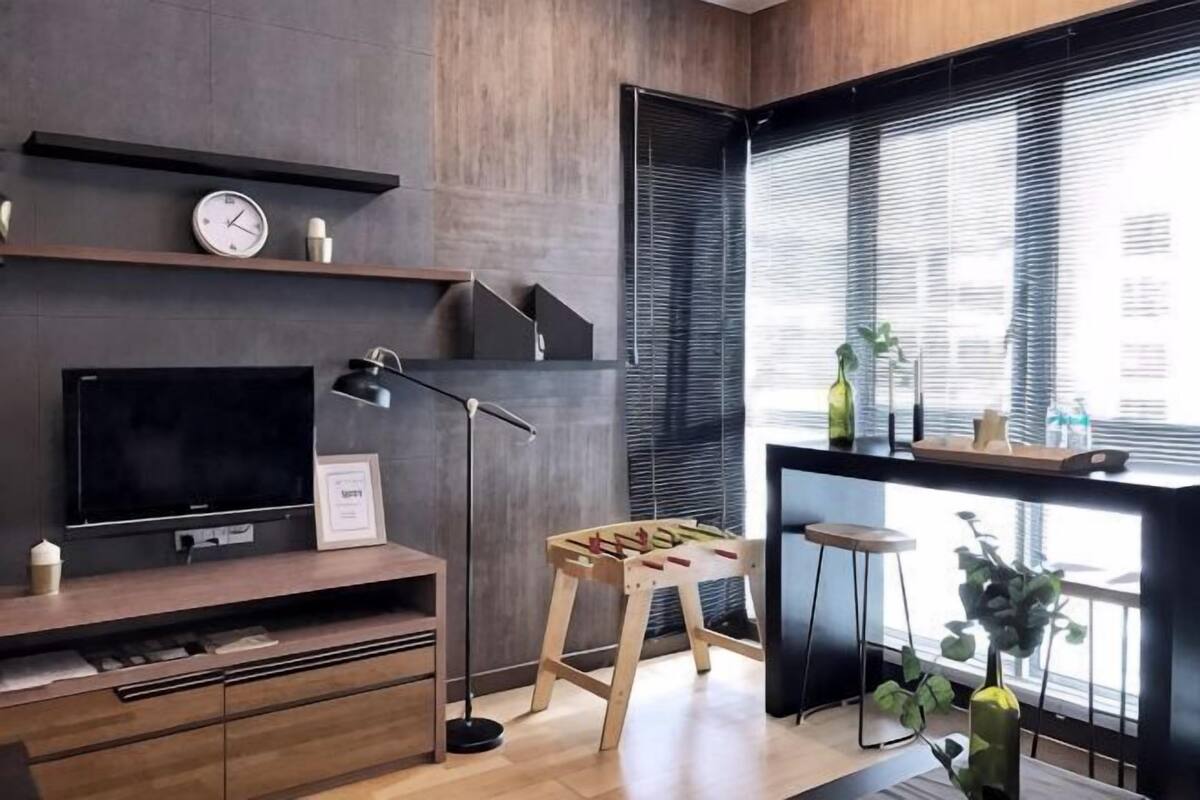 Ein modernes Wohnzimmer mit Flachbildfernseher, Stehlampe, Holzschemel und einem Fenster mit Jalousien.