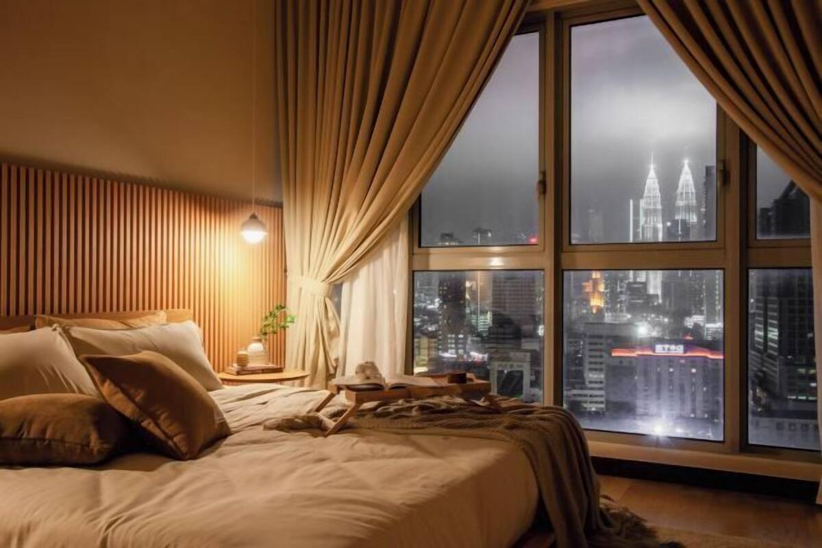 Ein Schlafzimmer mit großem Fenster, einem Bett mit Kissen, einem Nachttisch mit Lampe und Blick auf eine nächtliche Stadtlandschaft.
