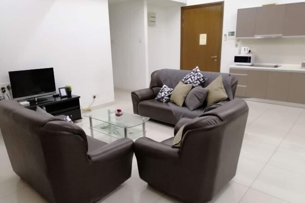 Ein modernes Wohnzimmer mit einer Couch, zwei Sesseln, einem Glastisch, einem Flachbildfernseher und einer Küchenzeile mit Schränken und Geräten.