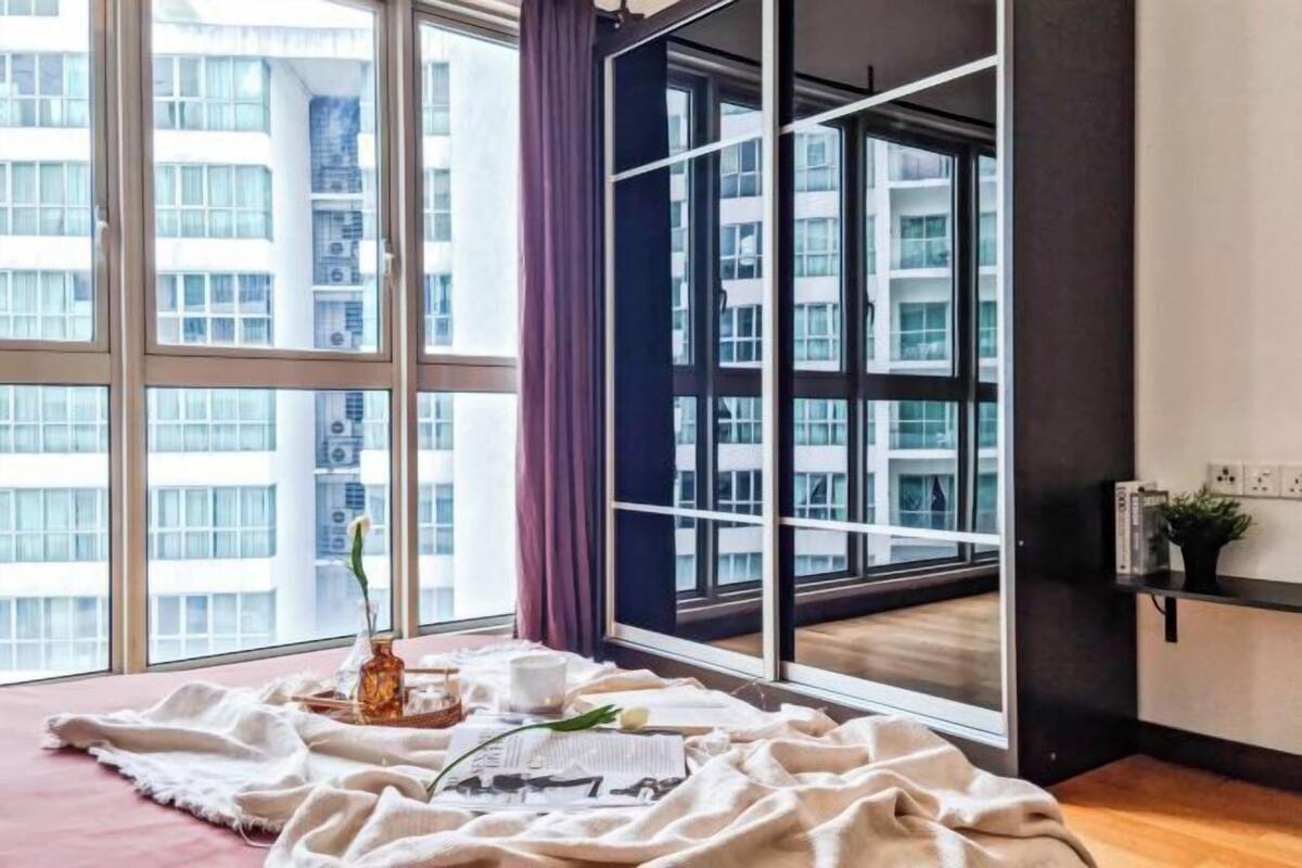 Ein Zimmer mit großen Fenstern, einem Bett mit weißer Decke, einer Vase mit Blumen und einem kleinen Tisch mit Tasse und Buch.