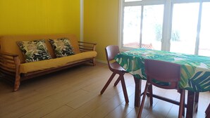 A room with a yellow wall, a wooden sofa with patterned cushions, a dining table with a green tablecloth, and a window with a view of greenery.
