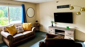 Un salon comprenant un canapé, un téléviseur, une petite table avec une plante et une lampe fixée au mur.