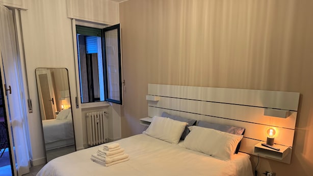 A neatly made bed with white linens and a stack of towels, a bedside table with a lamp, and a window with blinds. - Casetta Martesana - new one-bedroom apartment on the naviglio (Milano)