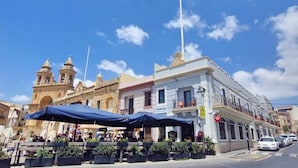 A street with outdoor seating, a building with a sign 'DUNCAN', and a historic building with bell towers. - Duncan guest house (Marsaxlokk)