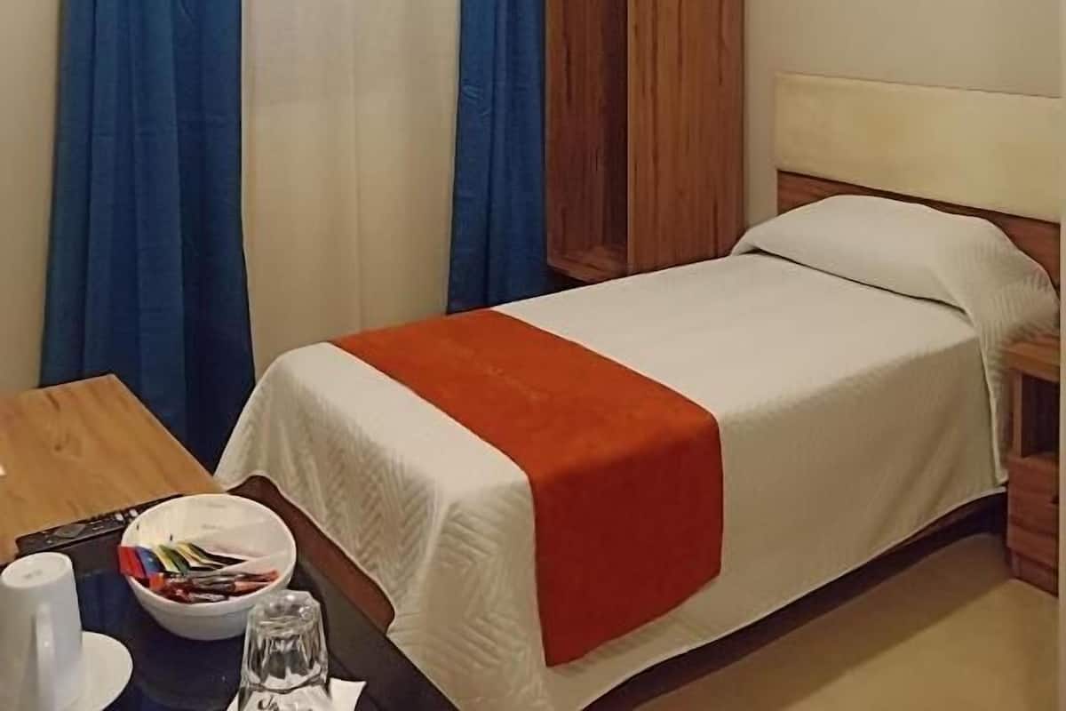 A hotel room with a single bed, a wooden wardrobe, a table with a cup and a bowl of fruit, and a curtain.