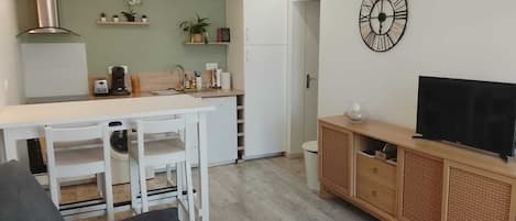 Uma sala de estar moderna com um sofá, uma mesa de centro, uma televisão sobre um móvel de madeira, e uma área de cozinha com uma geladeira branca, armários brancos e um relógio na parede.