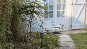 Polku johtaa talolle, jossa on valkoinen ovi ja ikkunat. Talon ympärillä on vehreää kasvillisuutta ja siellä on myös kaksi lyhtyä.
