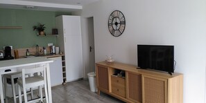A compact kitchen with a white refrigerator, a wooden cabinet with a TV, and a wall clock. - "Nid Serein" 2-room apartment with a garden and parking space; everything is within walking distance; close to downtown, the harbor, and the beach (Courseulles-sur-Mer)