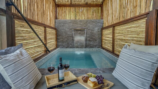A spa area with a hot tub, wooden walls, and a stone wall in the background. There are pillows and a tray with wine and snacks. - Kmar Villas (Maya Beach)