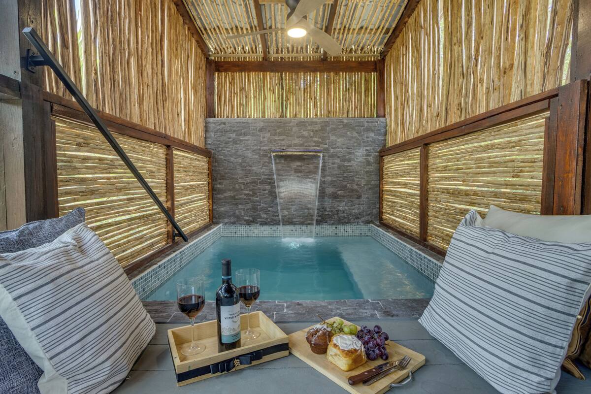 A spa area with a hot tub, wooden walls, and a stone wall in the background. There are pillows and a tray with wine and snacks.