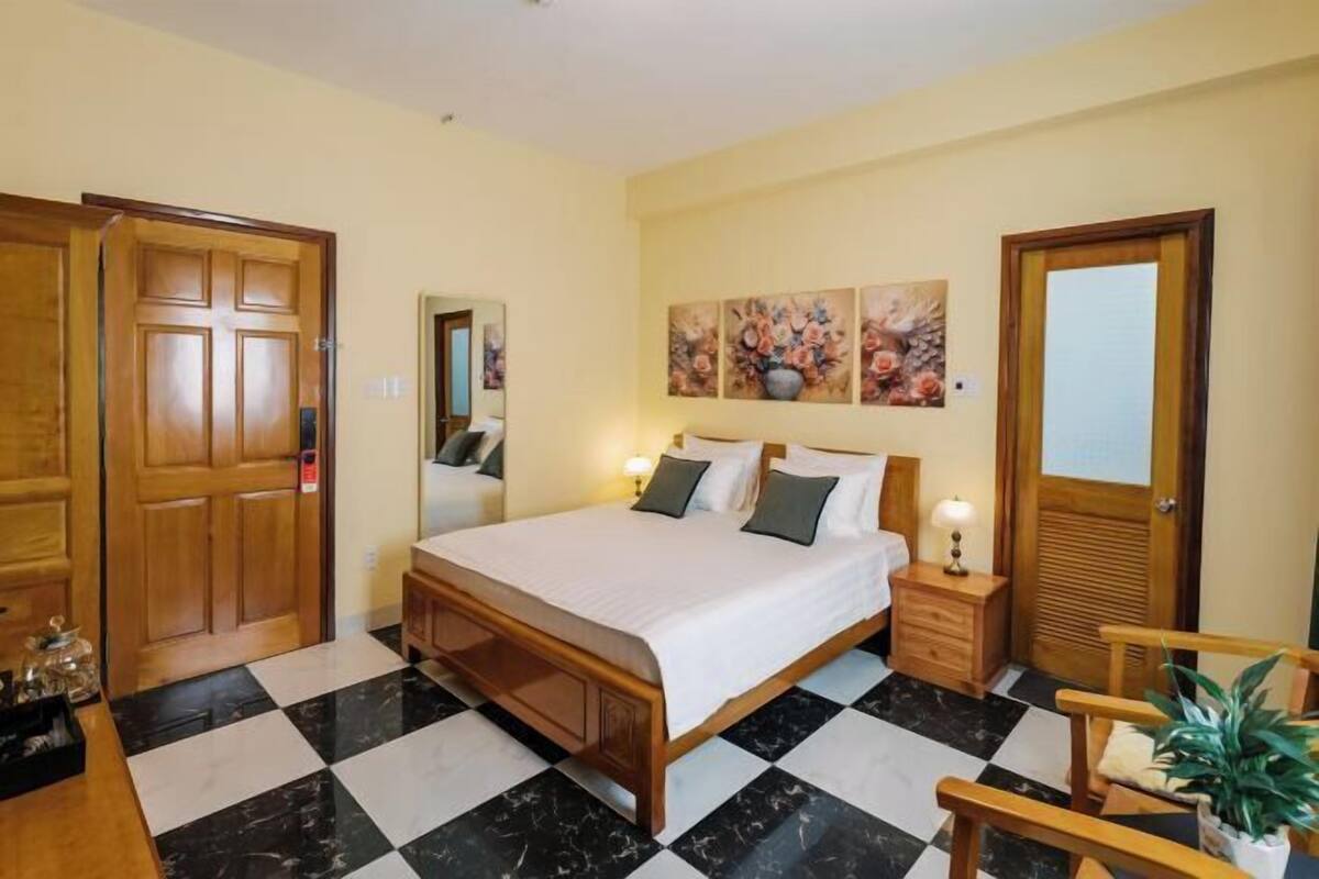 A hotel room with a checkered floor, a bed with pillows, a nightstand, a mirror, and wooden doors.