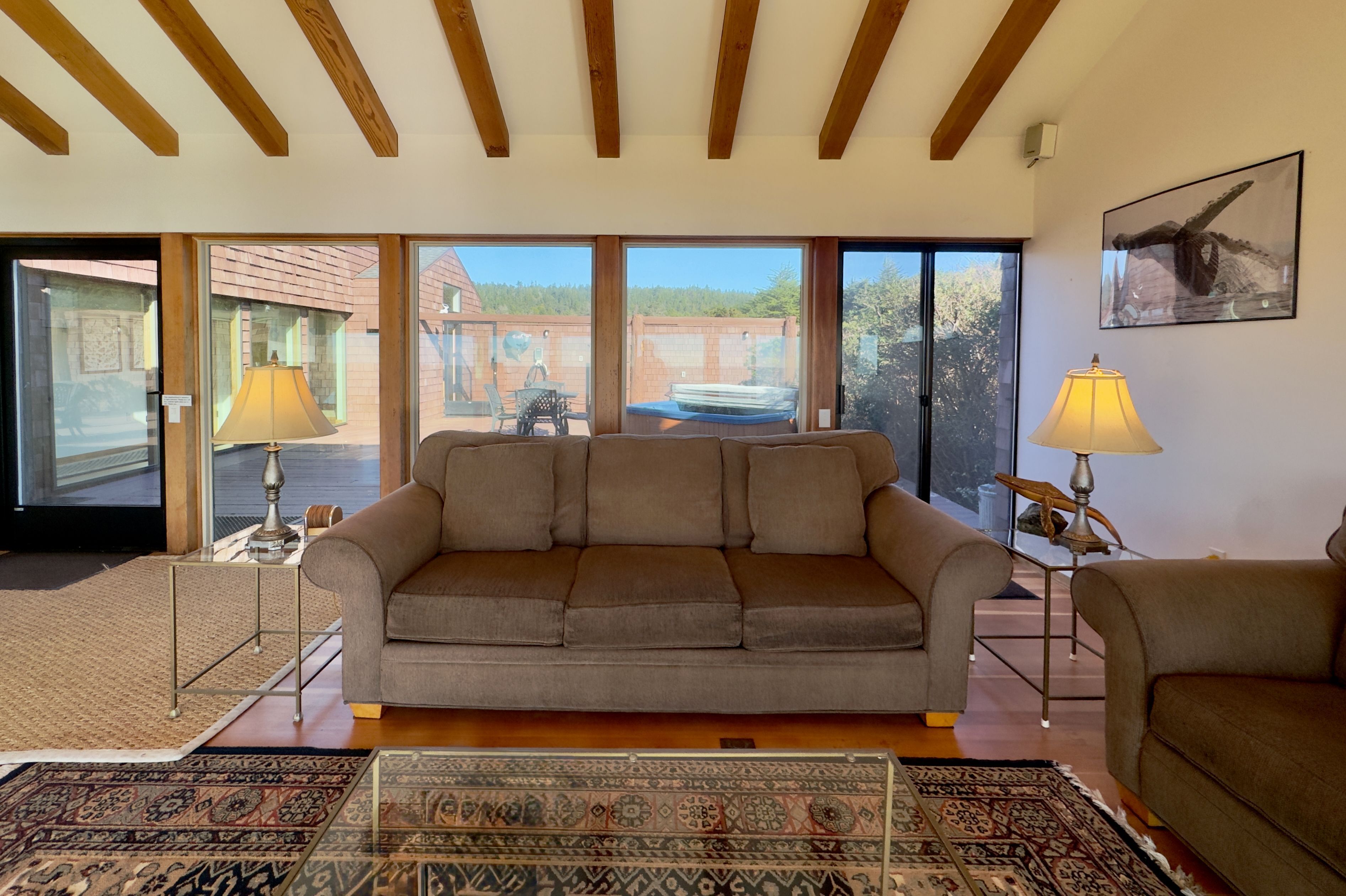 A living room with a beige sofa, two lamps, a patterned rug, and large windows offering a view of a backyard with a hot tub and patio furniture.