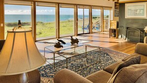 A spacious living room with large windows offering a view of the sea, a fireplace, and a coffee table with decorative items. - Seascape Oceanfront w/ hot tub, private courtyard (Sea Ranch)