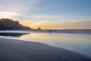 Une plage où l’on voit une personne portant une planche de surf, avec une eau calme et une falaise rocheuse en arrière-plan.