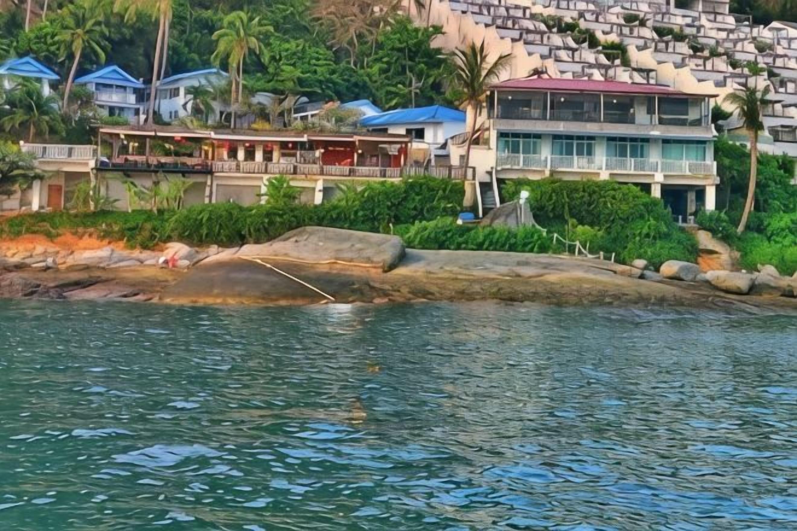 A waterfront property with a rocky shoreline, a building with a red roof, and a backdrop of hills with multiple houses.