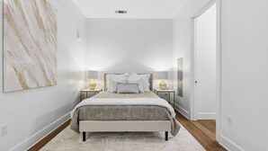 Un dormitorio con una cama, mesitas de noche, una lámpara, una obra de arte en la pared y una puerta.