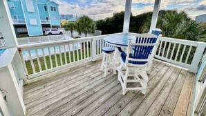 A wooden deck with a white railing, a table with blue chairs, and a view of a blue building in the background.