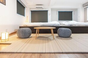 Una habitación de hotel moderna con cama, una mesita redonda, dos otomanes redondos, una lámpara de pie y una ventana con vistas.