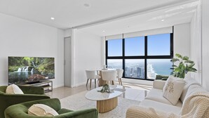 A modern living room with a large TV, green sofas, a white coffee table, and a view of the ocean through large windows. - Cozy 2 Bedroom 2 Bathroom Ocean View Unit - Q Stay (Surfers Paradise)