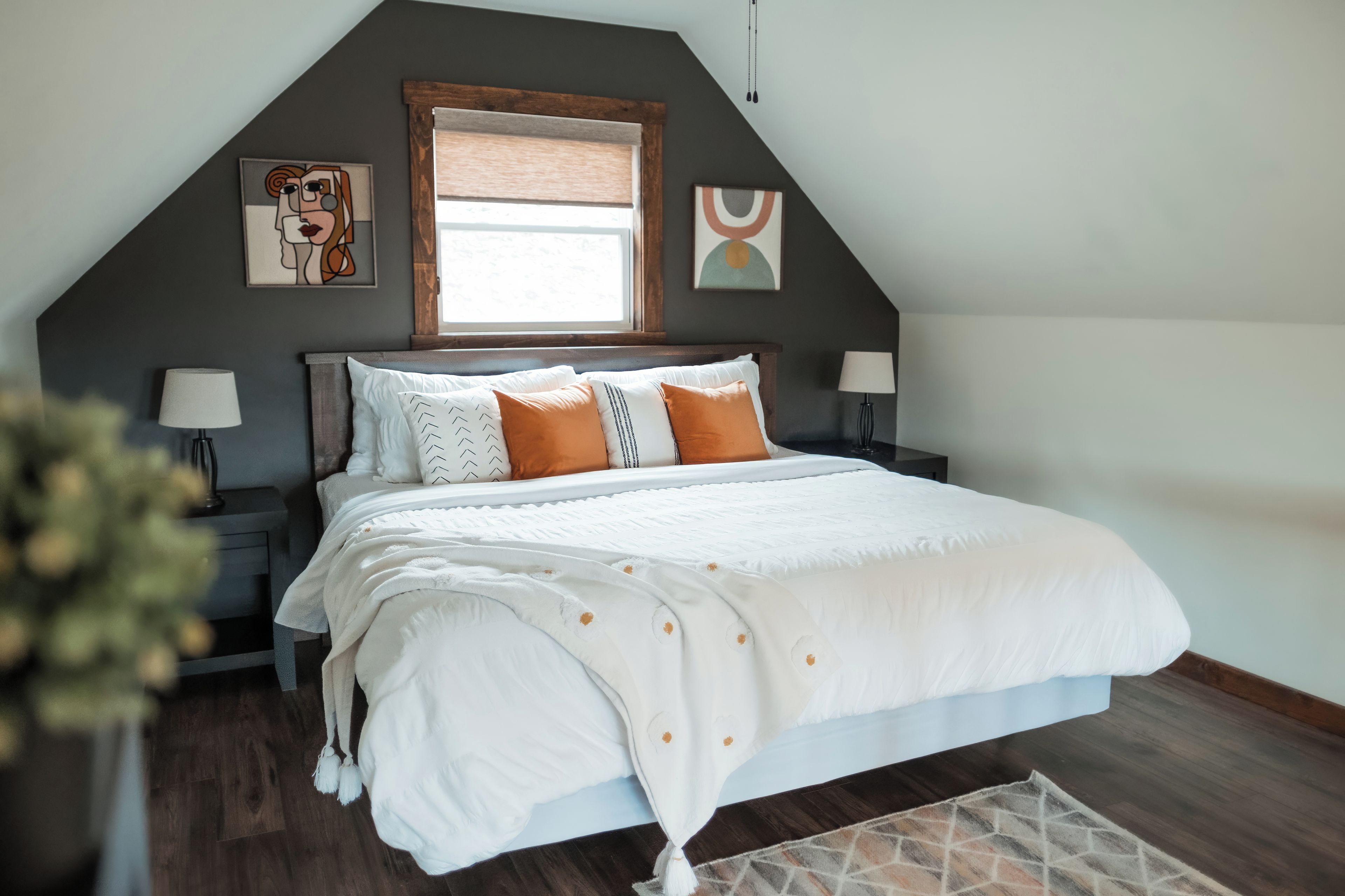 Une chambre à coucher avec un grand lit, deux tables de chevet avec des lampes, une fenêtre avec des stores et des œuvres d’art accrochées aux murs.