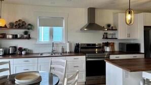 A modern kitchen with white cabinets, stainless steel appliances, a window with blinds, and a hanging pendant light.