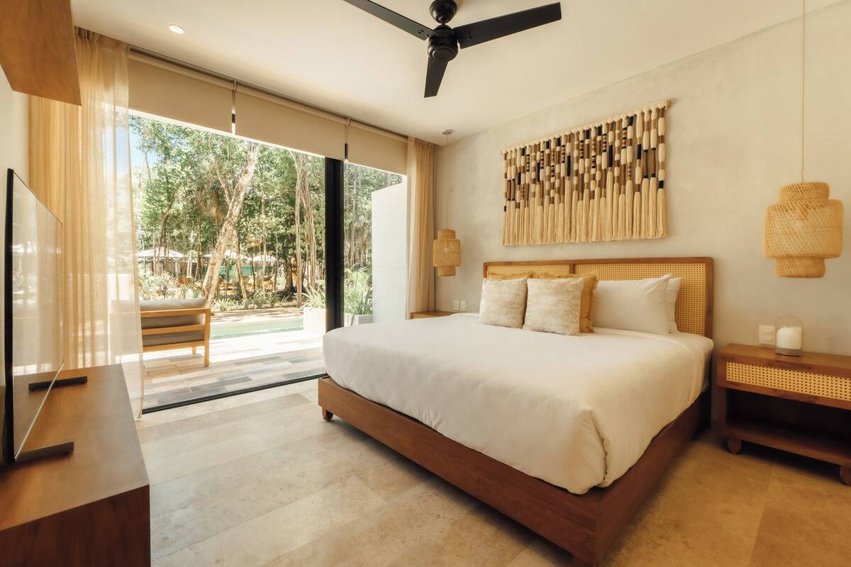 A spacious bedroom with a large bed, a ceiling fan, a TV, and a view of a pool and trees.