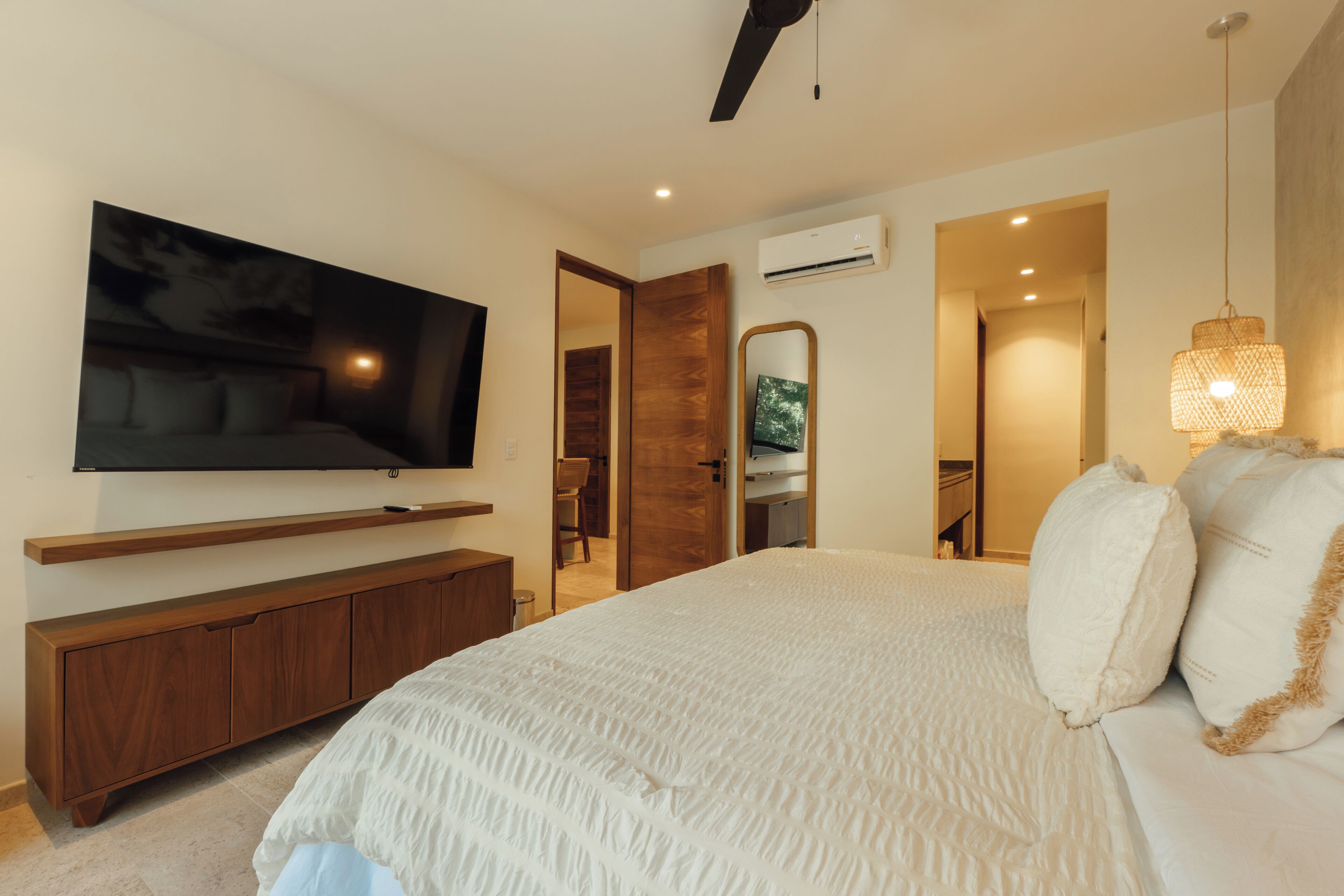 A modern bedroom with a large bed, a flat-screen TV mounted on the wall, a wooden cabinet, and a ceiling fan.