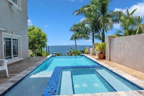 Ein rechteckiger Swimmingpool mit blauen Mosaikfliesen, umgeben von einer Steindeck, Palmen und Blick aufs Meer.