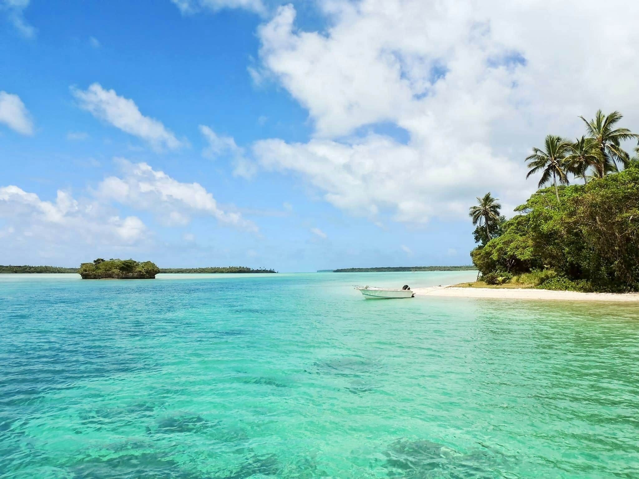 A clear turquoise sea with a boat anchored near a sandy beach, surrounded by lush greenery and palm trees.