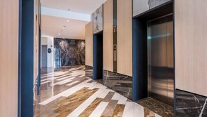 Un pasillo moderno con suelo de mármol, un ascensor y una pared con una gran obra de arte abstracta.