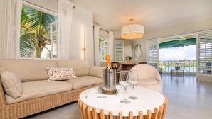 Un salon moderne avec un canapé beige, une table ronde en bois et une vue sur l’océan grâce à de grandes fenêtres.